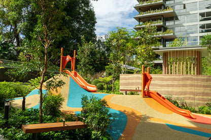 Two orange slides in a modern play area, set amidst greenery and colorful flooring, with a bright and inviting design.