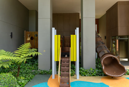 This image shows a modern playground area with a central pathway featuring yellow panels on either side, leading to a wooden play structure. A brown tube slide is visible on the right, and the space is surrounded by greenery, including ferns and ground cover. The area is partially enclosed with neutral-toned walls and columns, creating a sheltered play environment.