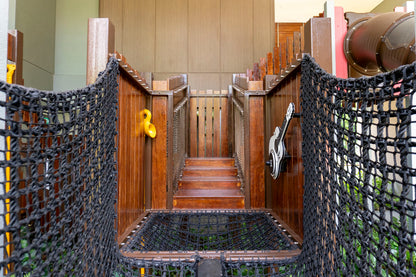 A wooden playground structure with a central staircase flanked by railings. The sides feature black netting for safety, resembling a rope bridge.